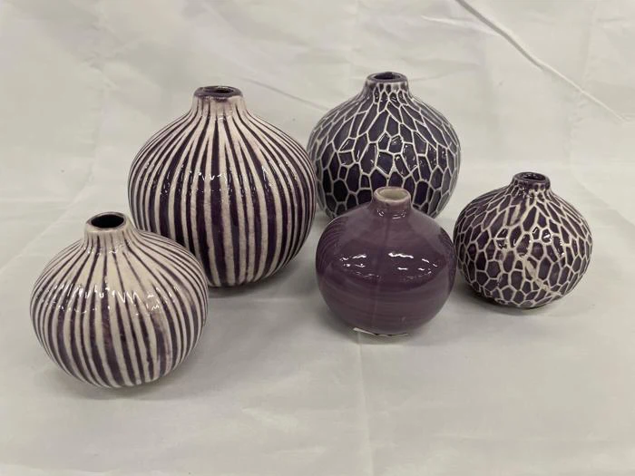Fem runde keramikvaser med smalle åbninger, med lodrette striber og geometriske mønstre i lilla og hvide nuancer, udstillet på en lys stofbaggrund.