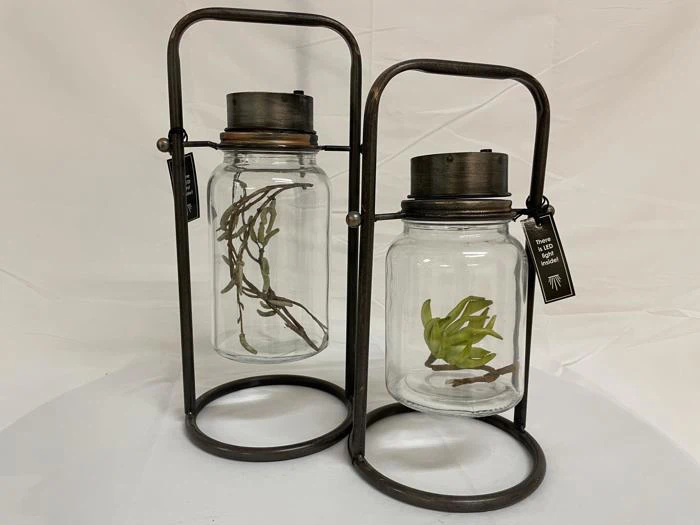 To glaskrukker med metallåg og stativ, hver med en kunstig plante, udstillet på en almindelig hvid baggrund.