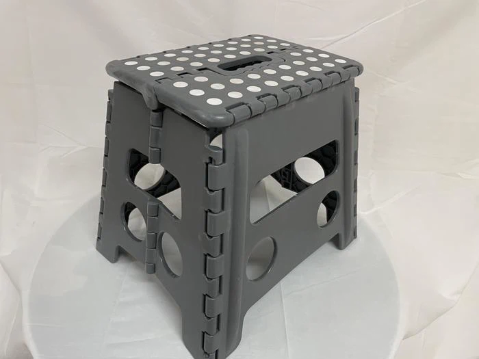 En sammenklappelig grå plastikskammel med hvide polkaprikker på toppen, placeret på en hvid stofbeklædt overflade.