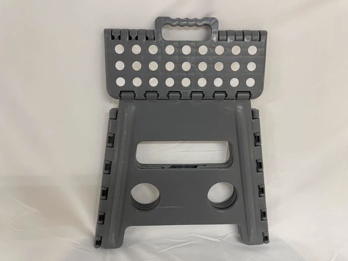 En grå, sammenklappelig skammel med håndtag og cirkulære huller i toppen og siderne, som står oprejst på en hvid flade.