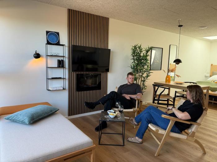 To personer sidder og snakker i en moderne, hyggelig stue med trægulv, et tv på væggen, et lille bord og minimalistisk indretning.