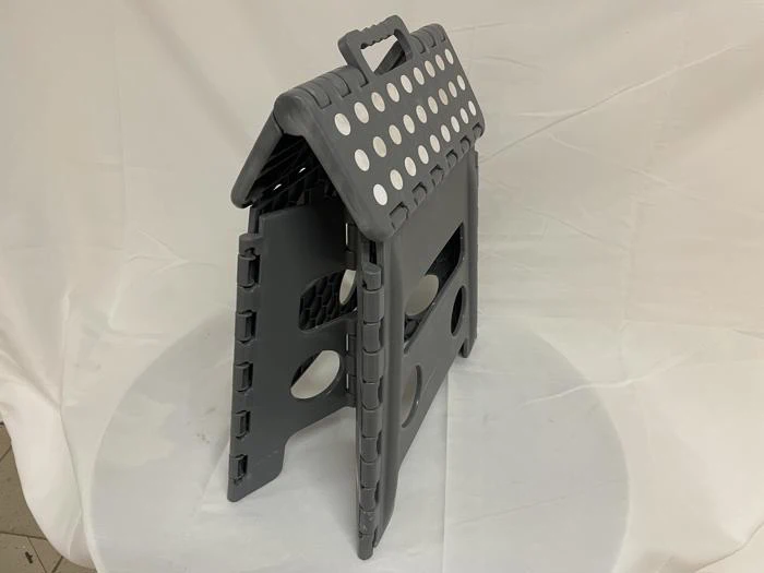 En grå, sammenklappelig plastikskammel med håndtag og cirkulære udskæringer, der står på en hvid stofbaggrund.