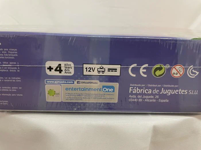 Nærbillede af en etiket til en legetøjskasse, der viser 4+ år, 12V, certificeringsmærker og producentoplysninger på spansk, med blå baggrund og noget tekst, der er delvist synlig.