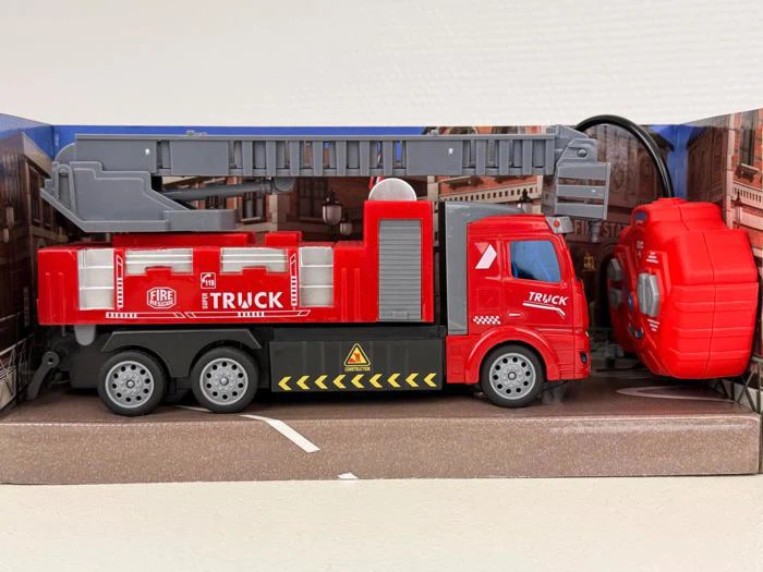 En rød legetøjsbrandbil med en udtrækkelig grå stige, pakket i en æske med bybaggrund. På siden af bilen står der FIRE TRUCK med hvide bogstaver.