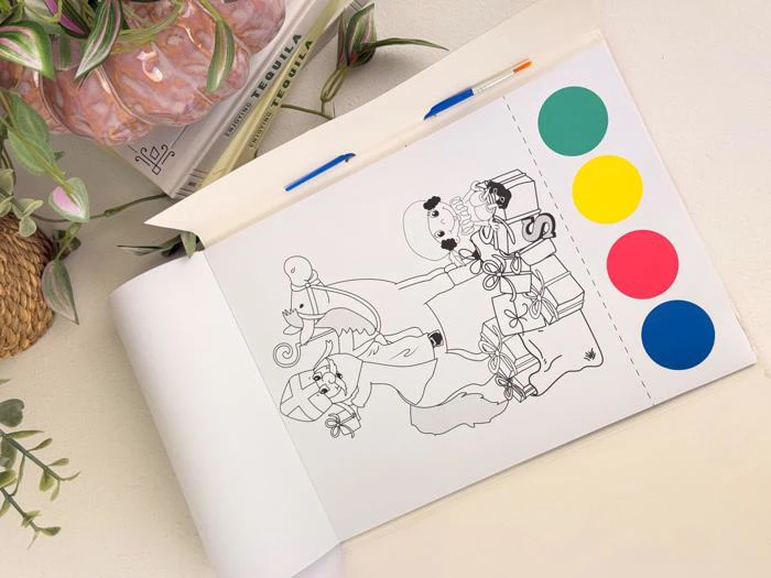 En malebogsside med julemanden, en elefant og gaver ved siden af fire farvecirkler (grøn, gul, rød, blå), en pen og nogle planter på et hvidt bord.