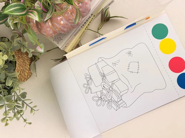 En åben malebog viser en plante i en syet potte med tre farvede cirkler (gul, grøn, rød) ved siden af. Potteplanter og kuglepenne står i nærheden på en hvid flade.