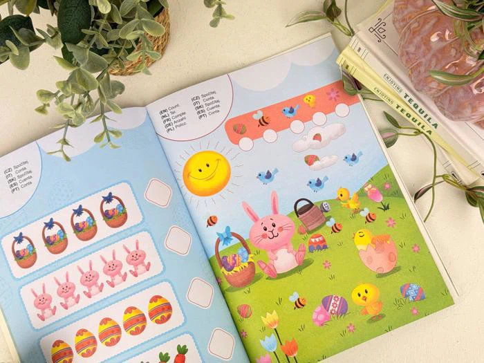 En åben aktivitetsbog viser farverige sider med påske-tema med æg, kurve, en lyserød kanin, fugle, bier, blomster og en smilende sol. Planter og bøger er i nærheden på en lys overflade.