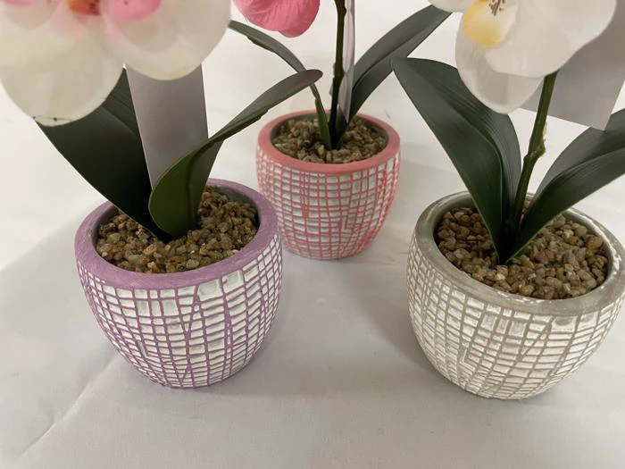 Tre små imiterede orkidéer i potter med stenjord, hver i en potte med struktur - en lilla, en rosa og en hvid - på en hvid overflade.