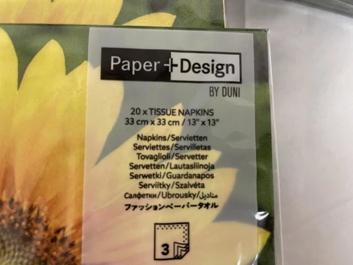 Et nærbillede af en servietpakke mærket Paper + Design by Duni med produktoplysninger på flere sprog og et solsikkeprint i baggrunden.