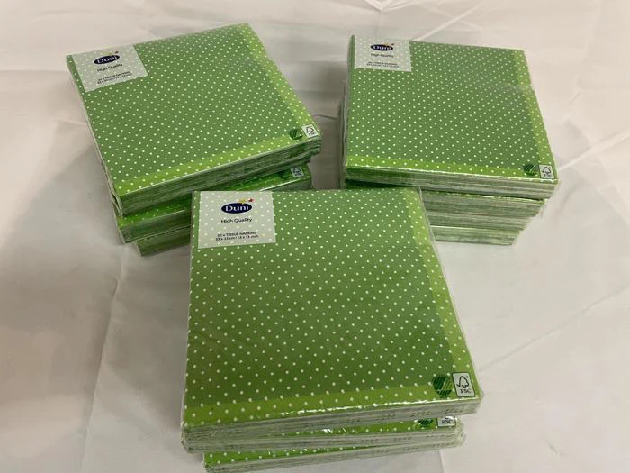 Fire stakke grønne papirservietter med hvide prikker er placeret på en hvid flade. Hver stak er pakket ind i plastemballage.