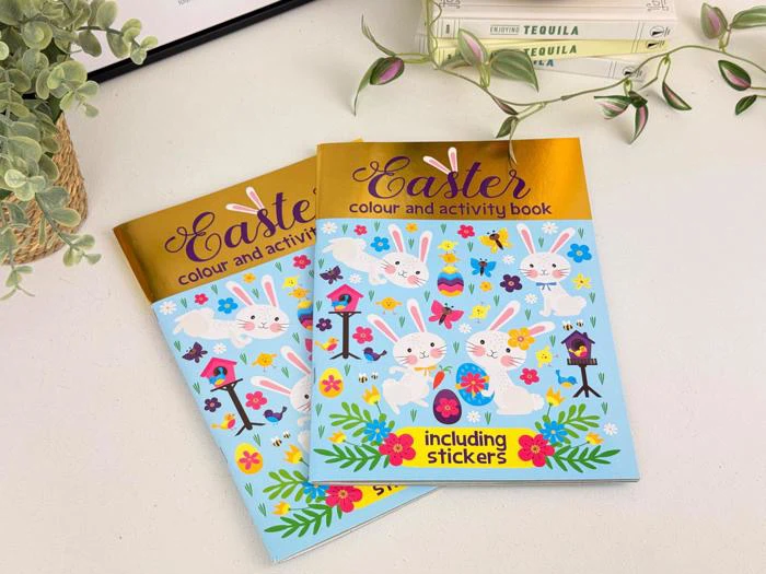 To farverige påskefarve- og aktivitetsbogsomslag med kaniner, blomster, fuglehuse og teksten inklusive klistermærker, placeret på et lyst skrivebord med dekorative planter.