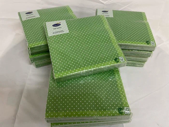 Fem pakker grønne papirservietter med hvide prikker, stablet på en hvid overflade. Hver pakke er pakket ind i plastik.