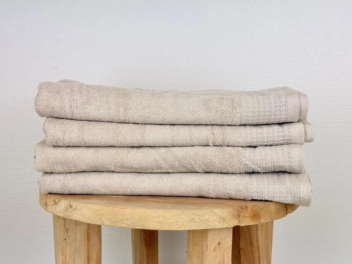 Fire pænt foldede beige håndklæder stablet på en rund træskammel mod en almindelig hvid struktureret væg.