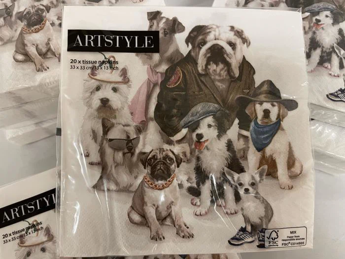 En pakke ARTSTYLE-servietter med illustrerede hunde i forskelligt tøj, bl.a. jakker, hatte og bandanaer, der poserer sammen i en gruppe.