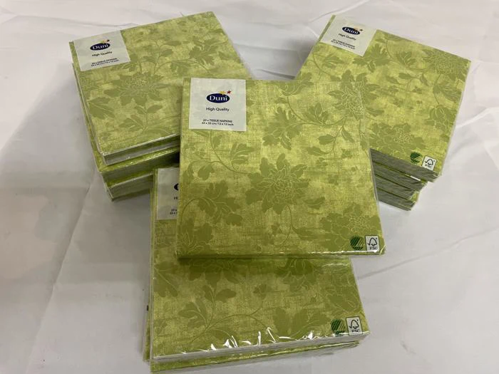 Stakke af grønne, blomstermønstrede papirservietter i plastemballage, arrangeret på en hvid flade. På etiketten står der "Duni High Quality.".