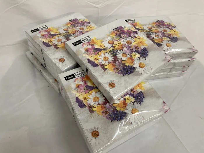 Stakke af indpakkede servietter med et blomstermotiv med margueritter og lilla og gule blomster på en hvid baggrund.