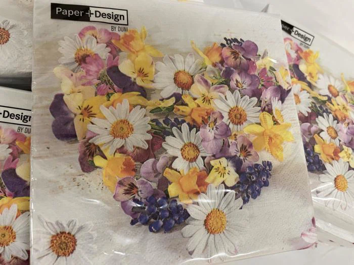En stak servietter med et hjerteformet blomsterdesign med margueritter, violer og gule blomster. På etiketten står der Paper+Design by Duni.