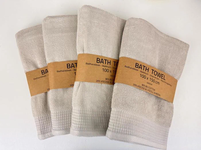Fire beige badehåndklæder, foldet og bundtet med brune papirbånd mærket BATH TOWEL 100 x 150 cm, er anbragt side om side på en hvid flade.