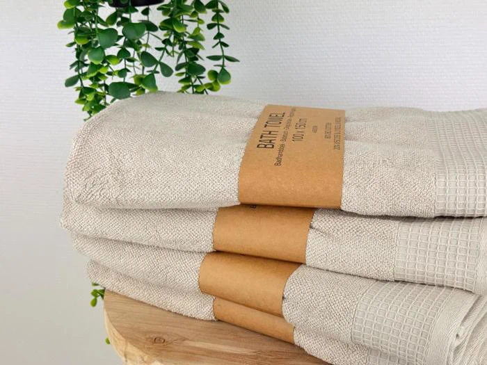 En stak beige badehåndklæder med brune papiretiketter ligger på en træplade med grønne hængende planteblade i baggrunden.