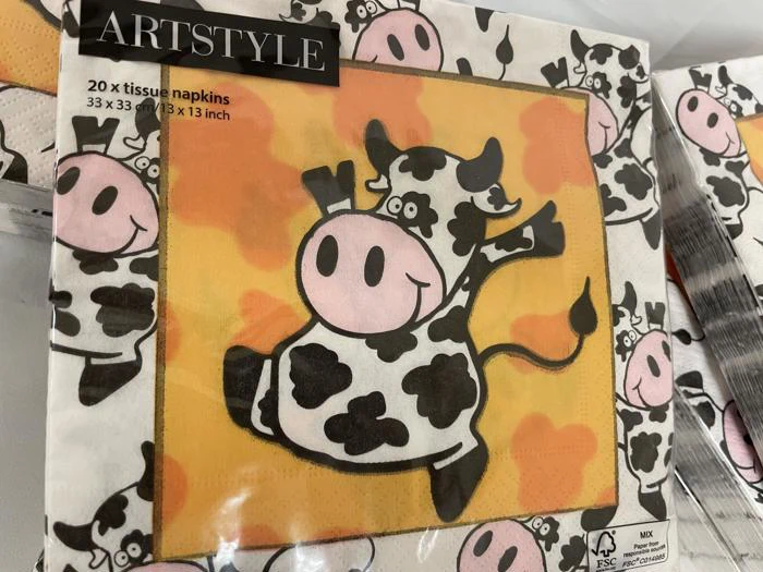 En pakke stofservietter dekoreret med en tegneserieko, der hopper på en orange og gul baggrund, omgivet af mindre ko-illustrationer.