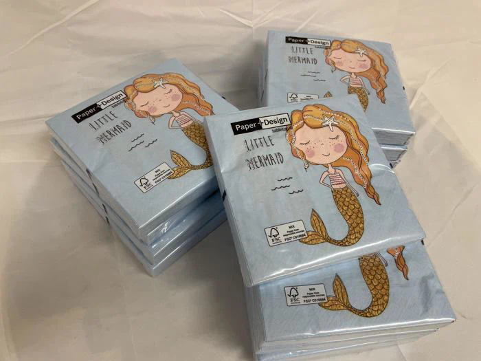 Flere pakker servietter med en illustration af en smilende havfrue og teksten Little Mermaid på en lyseblå baggrund er stablet på en hvid flade.