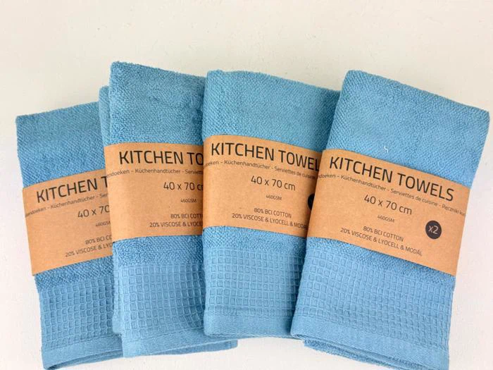 Fire foldede blå køkkenhåndklæder med strukturerede mønstre, hver indpakket i en brun etiket med teksten KITCHEN TOWELS, 40 x 70 cm, 80 % bomuld, 20 % viskose & lyocell.