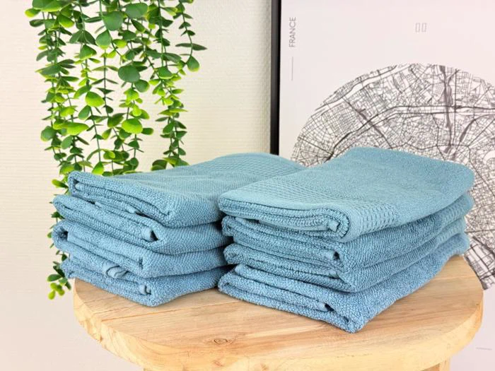 To pæne stabler af foldede blå håndklæder ligger på et træbord med en grøn bladplante og et sort-hvidt kort i baggrunden.