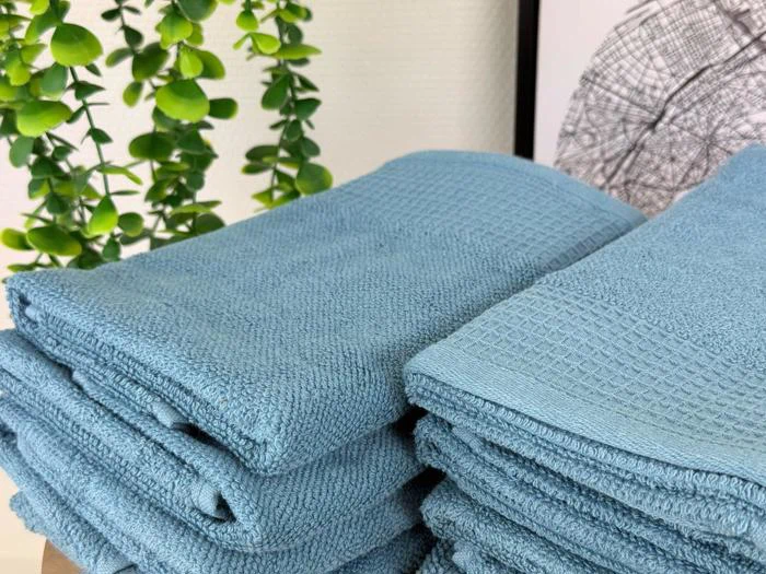 Stabler af foldede blå håndklæder er placeret på en flade med grønne bladplanter og et indrammet kunstværk i baggrunden.