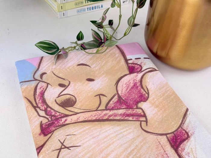 En notesbog med en skitse af Peter Plys på forsiden ligger på et skrivebord ved siden af en guldkrukke og en lille plante, og bøgerne er stablet i baggrunden.