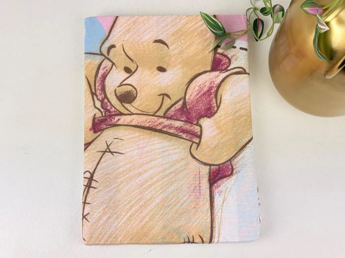 En stofbeklædt genstand med en skitse af Winnie the Pooh, der smiler, ved siden af en gylden vase med en grøn plante, på en lys overflade.