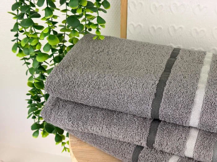 Tre pænt foldede grå håndklæder med hvide og mørke striber ligger stablet på en træplade ved siden af en grøn bladplante.
