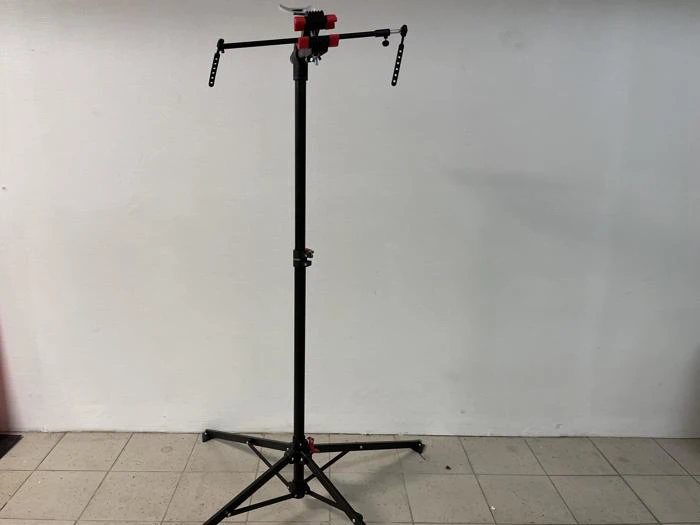 Et sort justerbart stativ med et rødt og sort armatur på toppen, der står på et flisegulv mod en almindelig hvid væg.