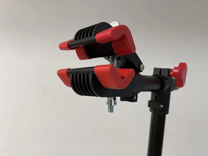 Et nærbillede af en sort og rød cykelreparationsklemme med polstrede kæber, monteret på en sort justerbar stang mod en almindelig baggrund.