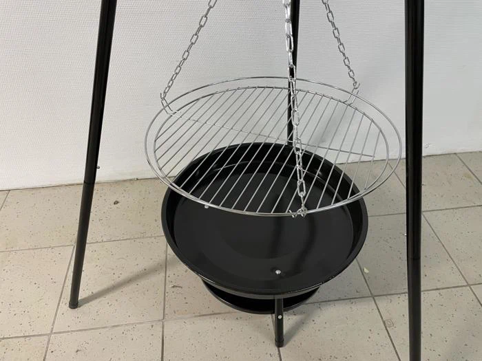 En trebenet metalgrill med en rund trådrist ophængt i kæder over en sort ildskål, der står på et flisegulv.