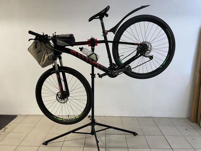 En sort og rød mountainbike med vandflaske og styrtaske er monteret på et reparationsstativ indendørs mod en hvid væg.