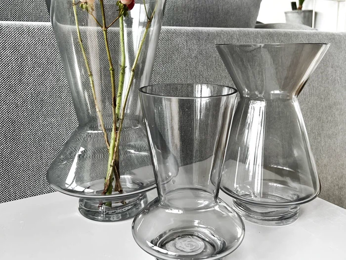 Tre tomme klare glasvaser med moderne geometriske former står på en hvid flade; i den ene vase er der et par høje blomsterstilke. I baggrunden er der grå, strukturerede møbler.