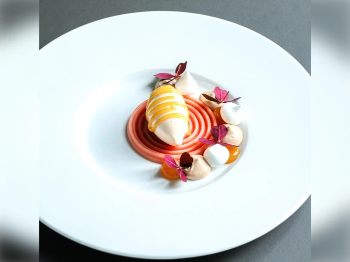 En dessert på tallerken med en central hvirvel af pink sauce, en quenelle toppet med gul sauce, klatter af fløde, orange gelé og spiselige lilla blade på en hvid tallerken.