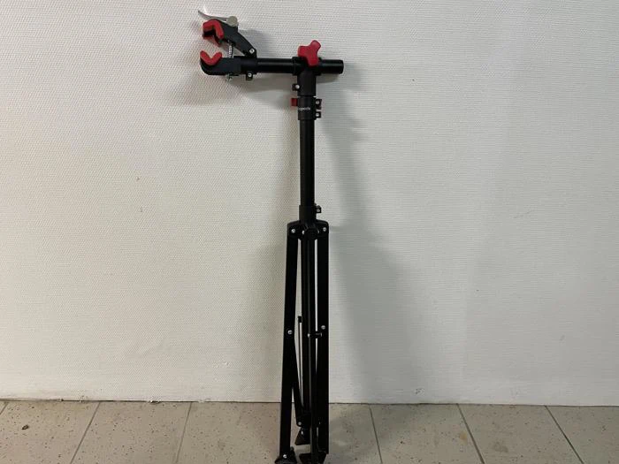 Et sort og rødt cykelreparationsstativ med en klemme, der står på et flisegulv mod en hvid struktureret væg.