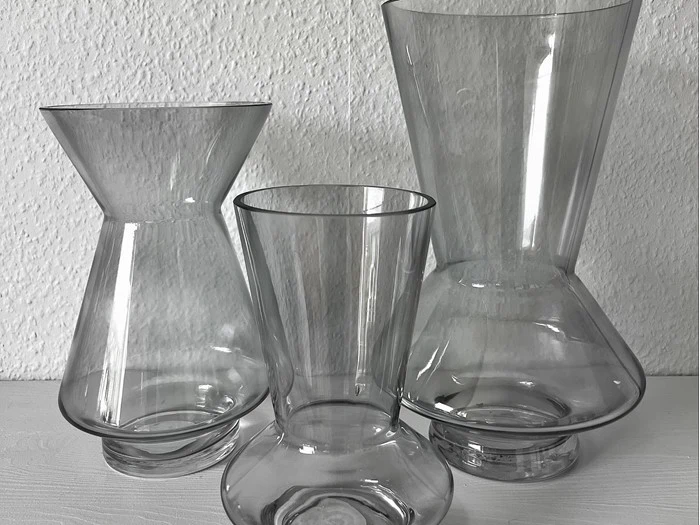 Tre tomme klare glasvaser i forskellige højder og unikke former er placeret på en hvid flade mod en struktureret hvid væg.
