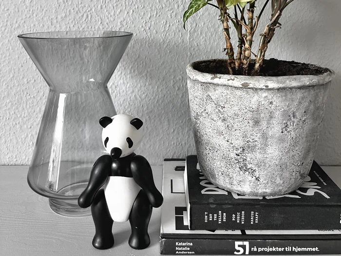 En glasvase, en lille pandafigur, en potteplante og stablede bøger er arrangeret på en lysegrå overflade mod en struktureret hvid væg.