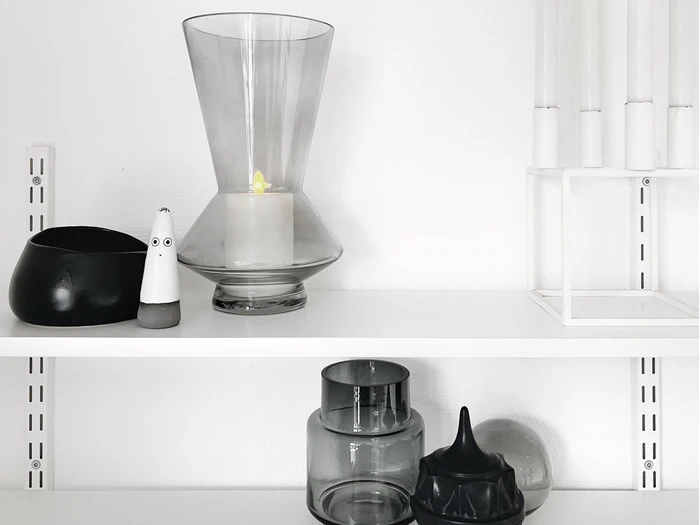 En minimalistisk hylde med dekorative ting: en stor glasvase med et stearinlys, en sort skål, en lille spøgelsesfigur, stablede glasbeholdere og hvide stearinlys i en holder.