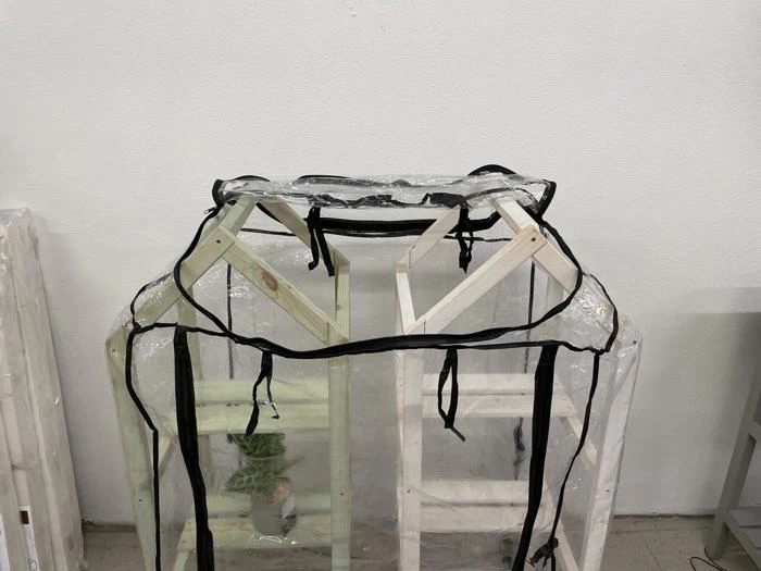 En lille planteramme af træ dækket af et drivhusovertræk af klar plast, delvist åbnet, står indendørs mod en hvid væg.