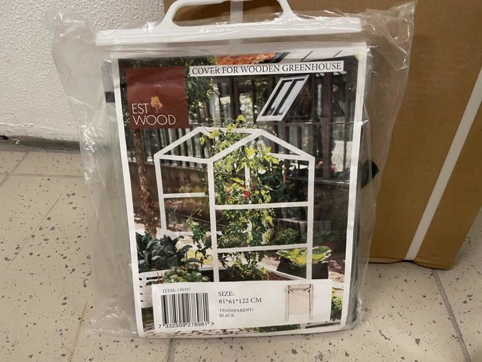 Et indpakket gennemsigtigt drivhuscover med et foto af et lille hvidt trædrivhus med planter indeni. På etiketten står målene 81x61x122 cm og mærket EST WOOD.