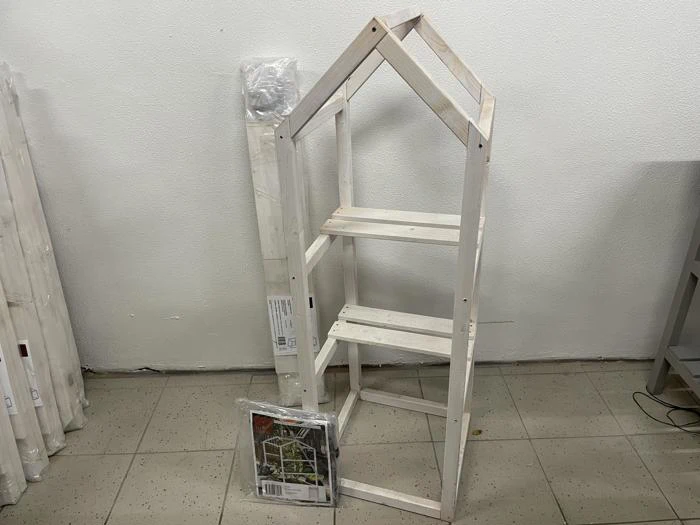 En lille, ufærdig træhylde formet som et hus står på et flisegulv med emballagematerialer og monteringsvejledning i nærheden.