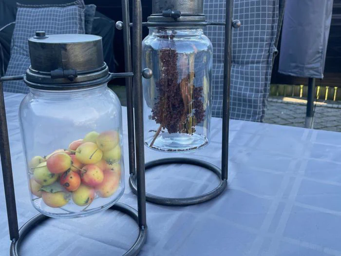 To glaskrukker på metalstativer; den ene fyldt med små gule og røde æbler, den anden med brunt tørret plantemateriale, på en blåternet dug udendørs.