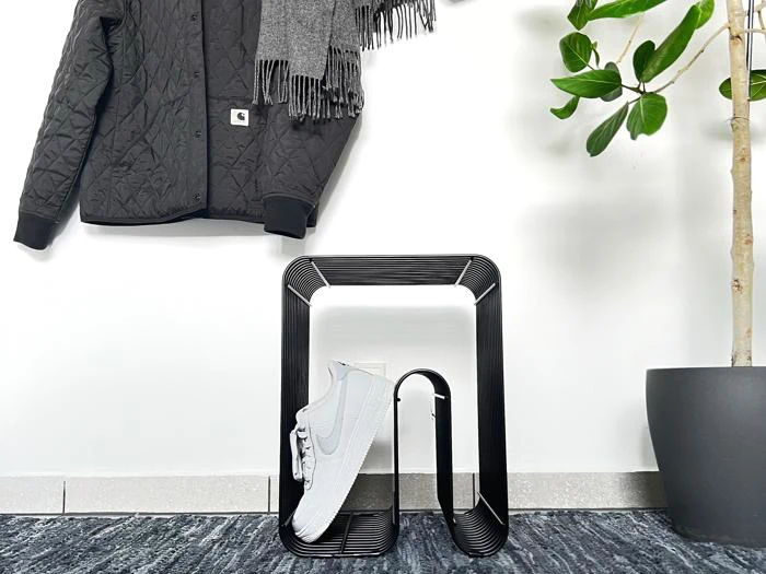 En quiltet sort jakke og et gråt tørklæde hænger på væggen over et moderne sort skostativ med en hvid sneaker; en potteplante står til højre.