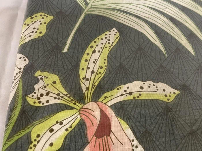 Nærbillede af mønstret stof med grønplettede orkidélignende blomster og palmeblade på en mørk, diamantmønstret baggrund.