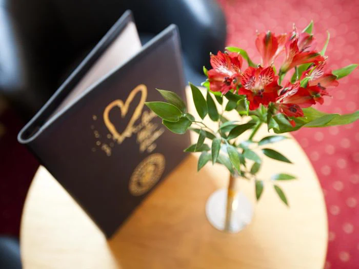 En lille vase med røde blomster står på et rundt træbord ved siden af et opretstående sort menukort med et guldfarvet hjertemotiv på forsiden.