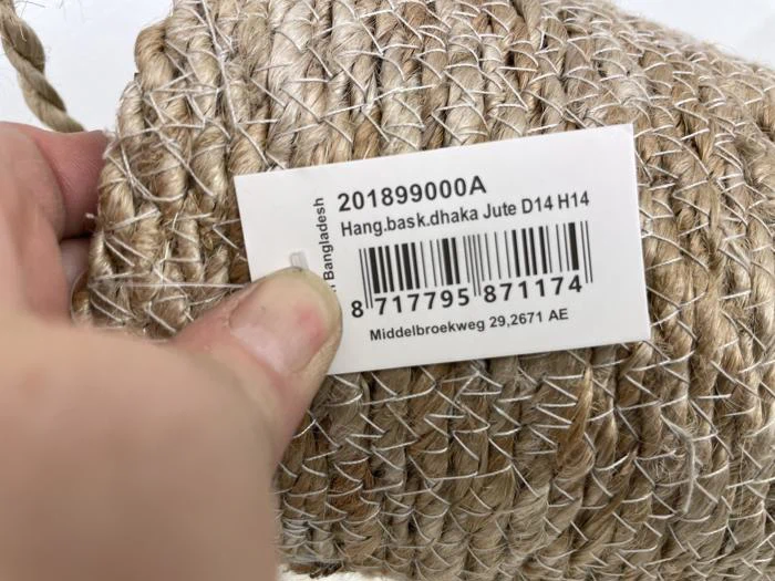 En hånd holder en rulle vævet jutereb med en hvid etiket, der viser en stregkode, produktoplysninger og en adresse i Holland.