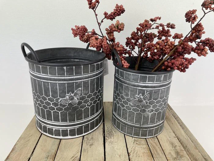 To dekorative metalspande med bi- og bikagedesign står på en træplade; den ene indeholder røde bærgrene, den anden er tom.